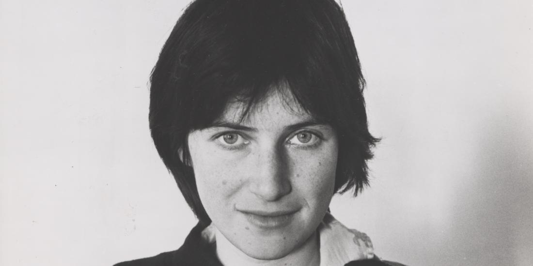 A black and white headshot of Chantal Akerman, who has one hand in her neck and stares straight into the camera, half-smiling.