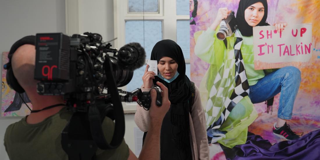 Young artist posing next to her work, a giant portrait of herself with the words 'Shut up, I'm talking'.