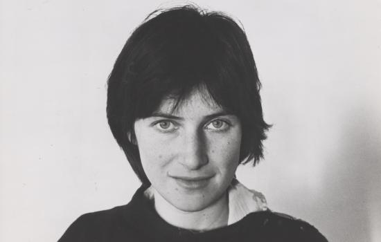 A black and white headshot of Chantal Akerman, who has one hand in her neck and stares straight into the camera, half-smiling.