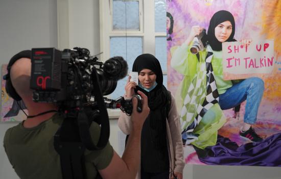 Young artist posing next to her work, a giant portrait of herself with the words 'Shut up, I'm talking'.