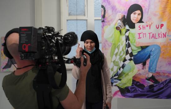 Young artist posing next to her work, a giant portrait of herself with the words 'Shut up, I'm talking'.