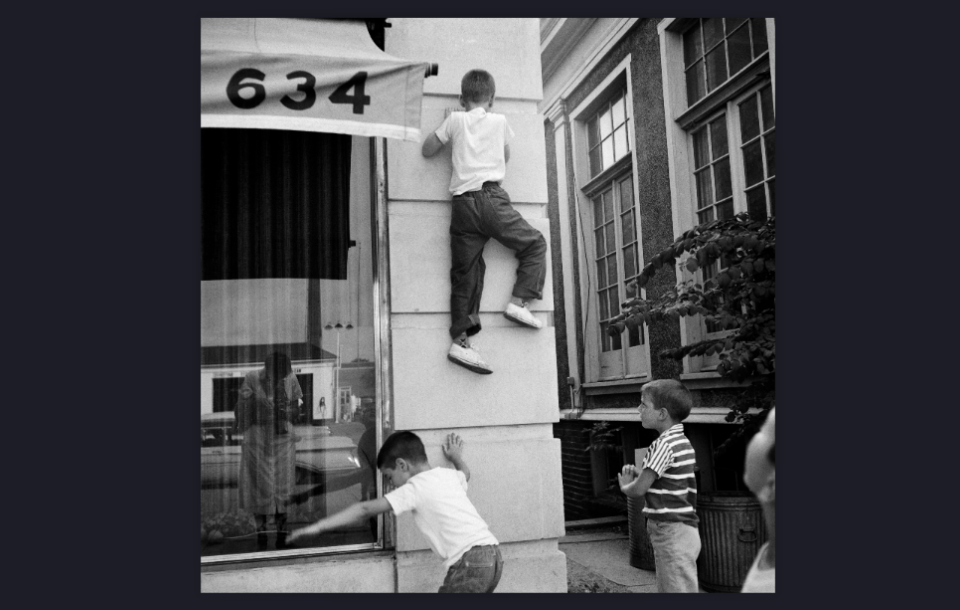 Zwart-witfoto van kinderen die een gevel opklimmen, in een winkelraam zie je de reflectie van de fotografe