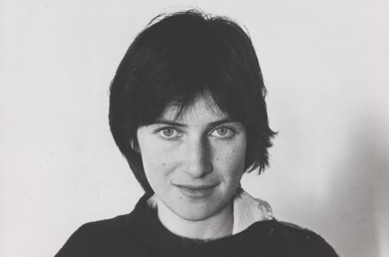 A black and white headshot of Chantal Akerman, who has one hand in her neck and stares straight into the camera, half-smiling.