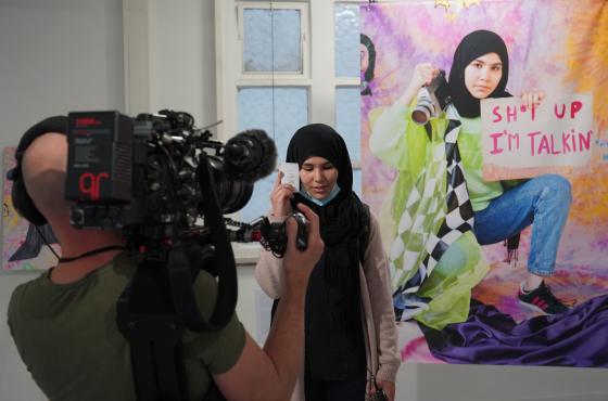 Young artist posing next to her work, a giant portrait of herself with the words 'Shut up, I'm talking'.